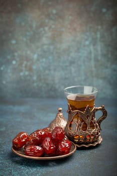 Breaking fasting with dried dates during Ramadan Kareem, Iftar meal with dates and Arab tea in traditional glass, angle view on rustic blue background. Muslim feast.