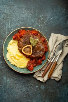 Traditional Italian dish Ossobuco all Milanese made with cut veal shank meat with vegetable tomato sauce served with corn polenta on ceramic plate top view on rustic stone background.