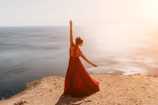 Side view a Young beautiful sensual woman in a red long dress posing on a rock high above the sea during sunrise. Girl on the nature on blue sky background. Fashion photo.