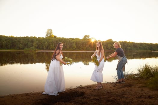 Three female girls in white sundresses and with flower wreaths on the riverbank at sunset. The feast of Ivan Kupala and national clothes and traditions of throwing wreath into the water