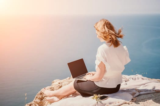 Successful business woman in yellow hat working on laptop by the sea. Pretty lady typing on computer at summer day outdoors. Freelance, travel and holidays concept.