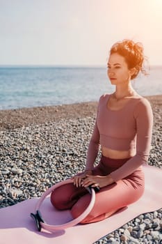 Middle aged well looking woman with black hair doing Pilates with the ring on the yoga mat near the sea on the pebble beach. Female fitness yoga concept. Healthy lifestyle, harmony and meditation.