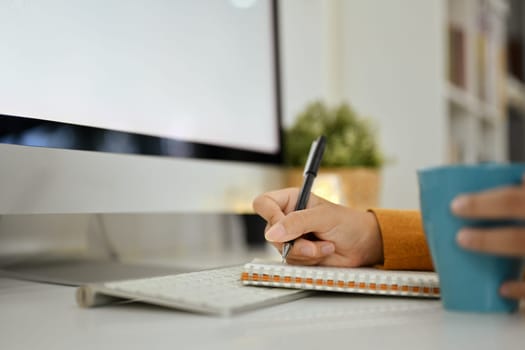 Side view of woman sitting in front of computer monitor and writing notes in personal daily planner.