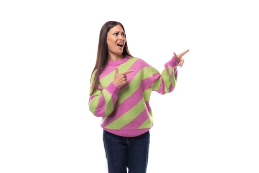 cute positive cheerful young european brunette woman dressed in a striped pink-green pullover points with her hand towards the empty space.