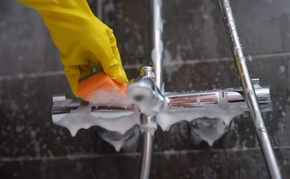 Cleaning lady in rubber glove washes faucet in bathroom with sponge with foam closeup. Cleaning company services concept