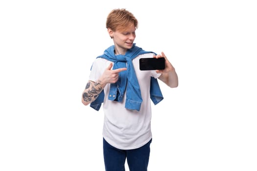 young emotional handsome slender red-haired man with a tattoo on his arms dressed in a stylish blue shirt shows ads on the phone.