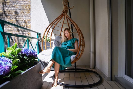 Woman in linen dress sits in hanging chair on a balcony