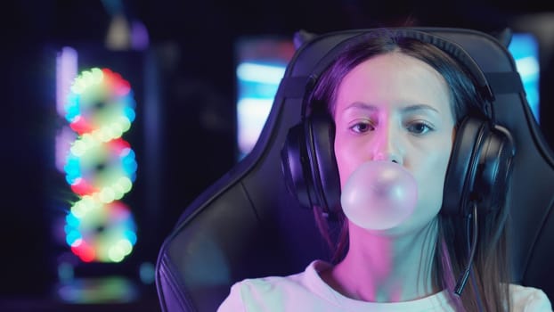 Young gamer woman sitting in a chair in gaming club and blowing a bubble gum, portrait