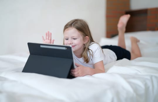 Little girl lying on bed and waving her hand at screen of tablet. Kids apps mobile concept