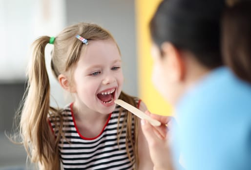 Pediatrician doctor examining throat of little girl with spatula. Chronic tonsillitis in children concept