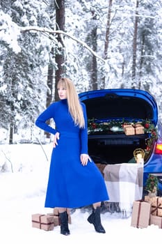Woman in winter snowy forest in blue dress next to blue car decorated with Christmas decor. Christmas and winter holidays concept