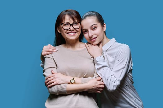 Portrait of middle aged mother and teenage daughter embracing together on blue studio background. Smiling happy mom and girl looking at camera. Family, lifestyle, relationship, mother's day concept