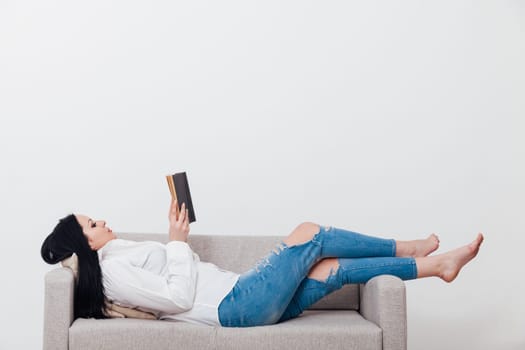 brunette woman reading a book on a gray sofa in a white room