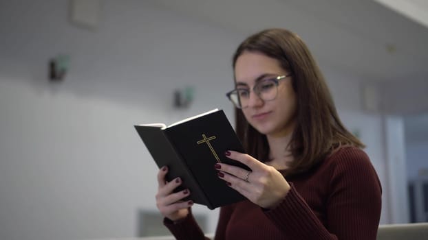 A young woman reads the Bible while sitting on a church bench. A Protestant girl with glasses reads the Bible in church. 4k