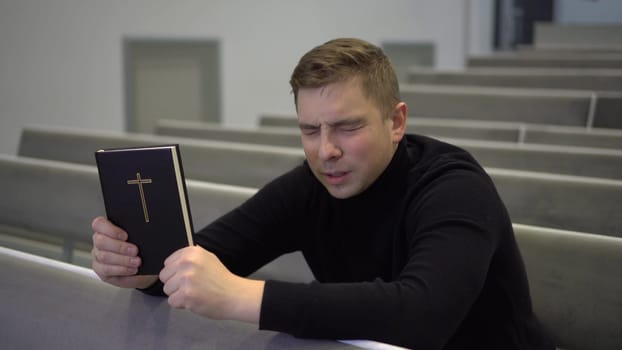 A young man prays holding a bible in his hands in a church. A Protestant man sits on a church bench and sincerely prays. 4k