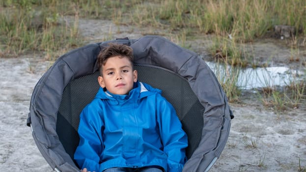 Portrait of a charming child boy in a tourist jacket sitting alone in a camping chair waiting for a hiker in nature with his family looking at the camera, High quality photo