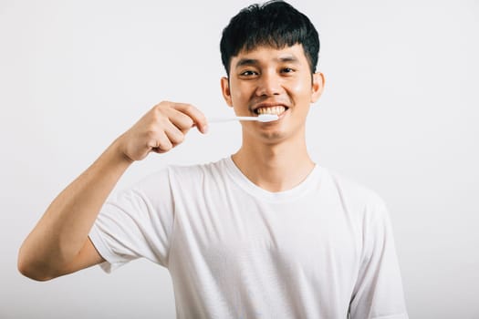 An Asian young man confidently brushes his teeth in the morning, promoting dental health. Portrait of a happy Thai teen with a toothbrush in a studio shot isolated on a white background.