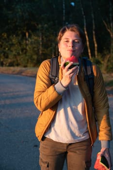 Young Caucasian blonde millennial woman looking like a guy eating watermelon in the sunny golden hour at sunset, wearing light jacket with backpack, High quality photo