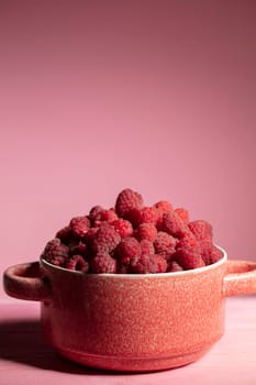 Vertically raspberries in a clay bowl on a pink background.