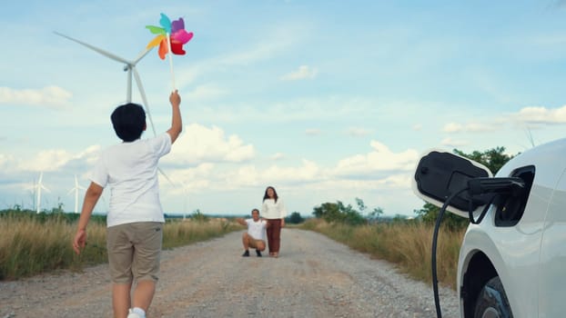 Concept of progressive happy family enjoying their time at wind farm with electric vehicle. Electric vehicle driven by clean renewable energy from wind turbine generator for charging station.