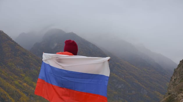 The flag of Russia flutters beautifully behind a woman's back against the backdrop of mountains on a cloudy day. Close-up rear view. Russian Independence Day.