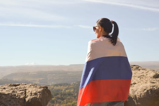 A girl tourist with a Russian flag on her shoulders stands on the top of a mountain and enjoys the beautiful views of the Caucasus Mountains