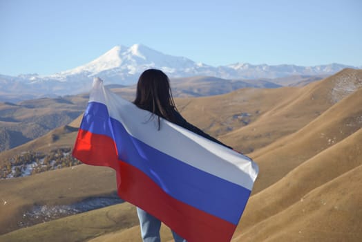 A young brunette woman stands against the backdrop of the snow-capped Mount Elbrus, looking at the beauty of the mountains, holding a Russian flag in her hands. Tricolor against the backdrop of snow-capped Mount Elbrus.