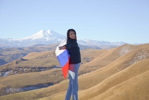 A young brunette woman stands against the backdrop of the snow-capped Mount Elbrus, looking at the camera, a Russian flag covers her shoulders. Tricolor against the backdrop of snow-capped Mount Elbrus.