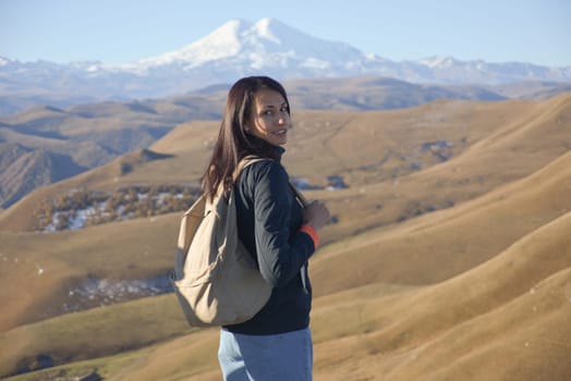 A young brunette woman with a backpack stands against the backdrop of snow-capped Mount Elbrus and looks at the camera
