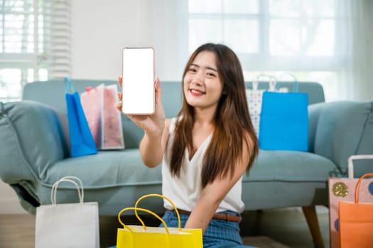 Excited Asian buying woman smiling showing mobile phone with many shopping bags at home in living room, Happy female online shopping payment show smartphone blank screen, Black Friday