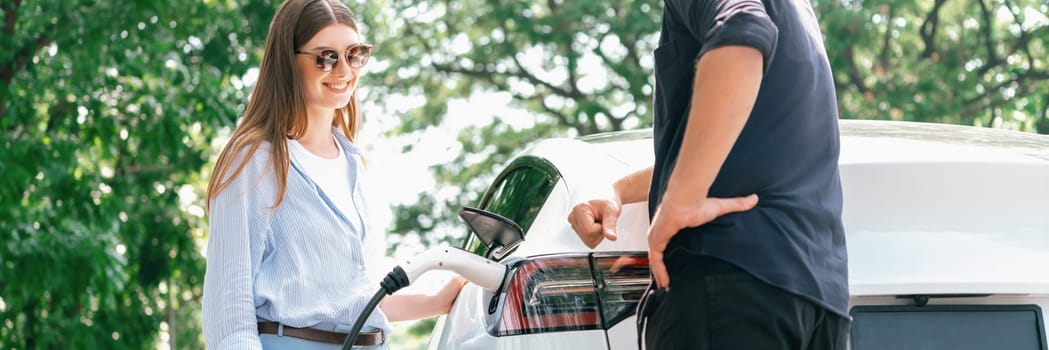 Focused EV car recharging electricity for battery on blurred background of lovey couple ruing their road trip travel by eco friendly electric car in national park and greenery forest on holiday. Exalt