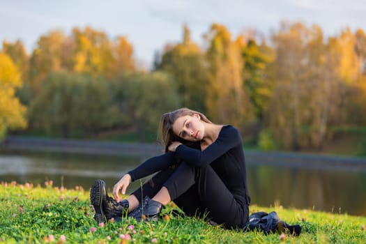 A beautiful girl poses while sitting on the grass by a pond in an autumn park