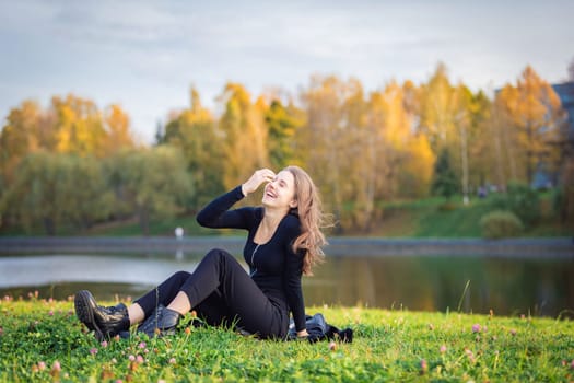 A beautiful girl poses while sitting on the grass by a pond in an autumn park