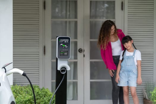 Happy little young girl learn about eco-friendly and energy sustainability as she help her mother recharge electric vehicle from home EV charging station. EV car and modern family concept. Synchronos