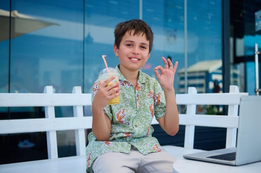 Handsome schoolboy in summer shirt, holds a glass of freshy squeezed orange juice, shows OK hand sign, confidently looks at camera, sitting at table with a laptop in a cafe, studying online outdoor