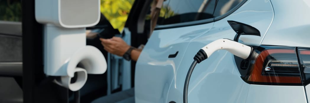 Young man recharge electric car's battery from charging station in outdoor green city park. Rechargeable EV car for sustainable environmental friendly urban travel. Panorama Expedient