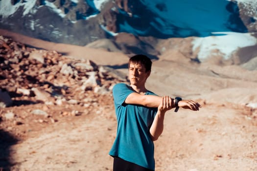 A young man does a warm-up before a trail and canyon race in the snow-capped mountains. The runner trains outdoor, does exercises in beautiful area.