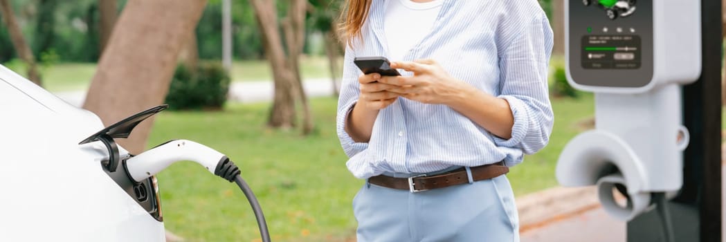 Young woman using smartphone online banking application to pay for electric car battery charging from EV charging station during vacation road trip at national park or summer forest. Panorama Exalt