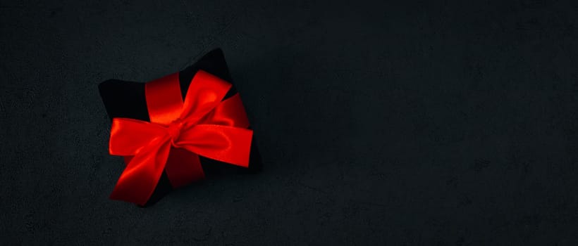 A black small velor gift pillow with a red ribbon tied in a bow lies on the left on a black background with space for text on the right, close-up side view. Christmas and black friday concept.