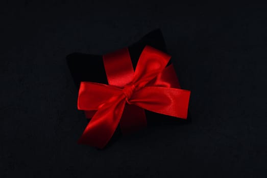 Black small velor gift pillow with a red ribbon tied in a bow lies in the middle on a black background, close-up side view. Christmas and black friday concept.