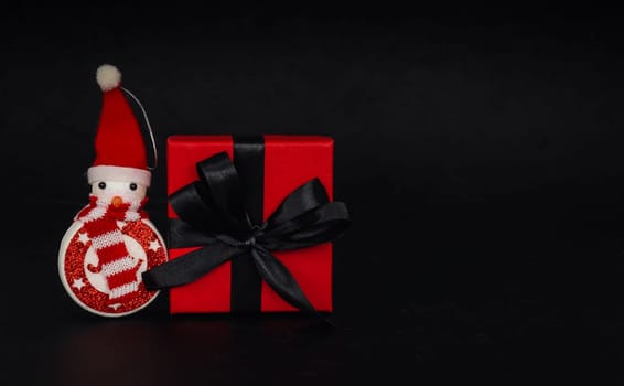 One red gift box and decorative snowman lies on the left against a black background with space for text on the right, close-up side view. Concept black friday, christmas.