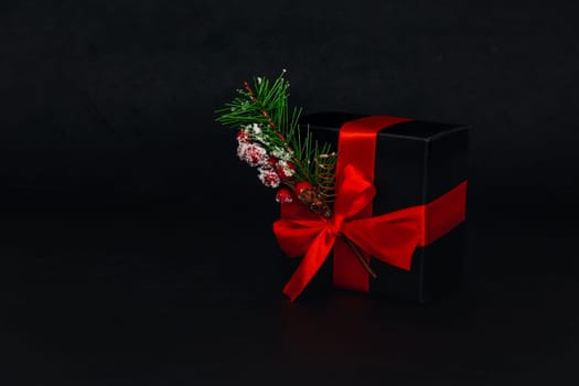 One red small gift box with a spruce branch and a black ribbon tied with a bow is on the right against a black background with space for text on the left, close-up side view. Christmas and black friday concept.