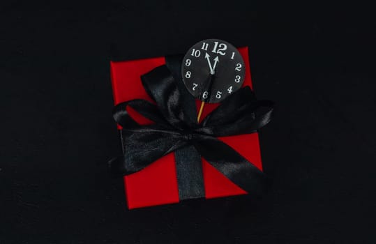 One small red gift box with a black ribbon and a paper clock in the middle on a black background with space for text, close-up top view. Christmas and black friday concept.