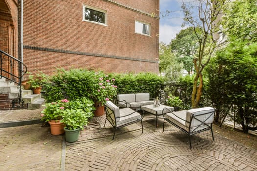 a patio with chairs and plants on the ground in front of a red brick building, surrounded by green trees