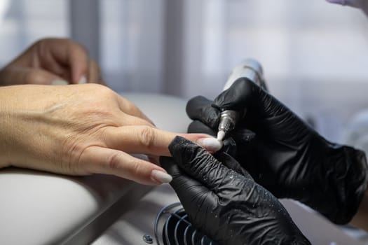 Manicurist at work. Professional manicurist removes old worn nail design from nails of client using modern electric drill. Preparing fingernails for making fresh manicure and applying new nail design