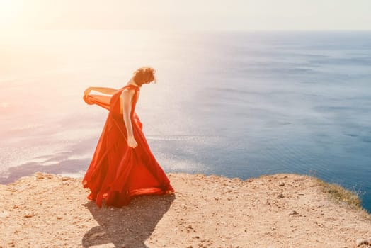 Side view a Young beautiful sensual woman in a red long dress posing on a rock high above the sea during sunrise. Girl on the nature on blue sky background. Fashion photo.