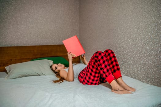 Young brunette woman works and studies from home on a laptop, while lying on her bed in red checkered pants. Balancing work and studies at home