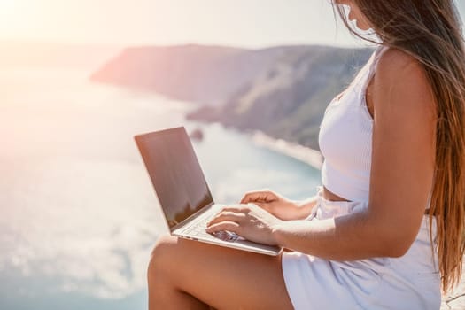 Successful business woman in yellow hat working on laptop by the sea. Pretty lady typing on computer at summer day outdoors. Freelance, travel and holidays concept.