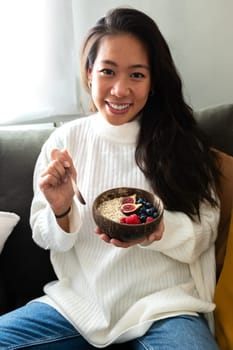 Happy young Asian woman smiling looking at camera showing bowl of healthy breakfast oats with fruit sitting on couch. Vertical image. Healthy lifestyle concept.