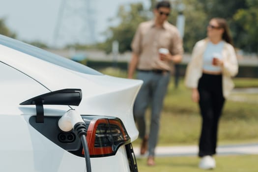 Young couple with coffee cup, recharge electric car's battery from EV charging station in green city park. Sustainable and eco friendly EV car with urban and shopping lifestyle. Expedient